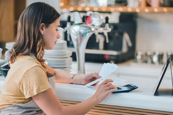 A picture of a young female business owner using a digital tablet and calculator while reviewing paperwork.